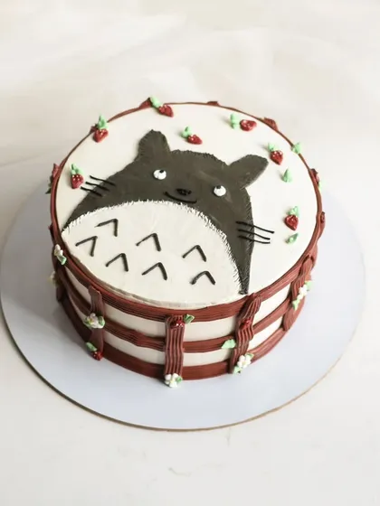 A cute hand-painted cake featuring Totoro from Studio Ghibli. The design is framed with a piped border and tiny strawberries, making it a whimsical treat for an anime fan.
