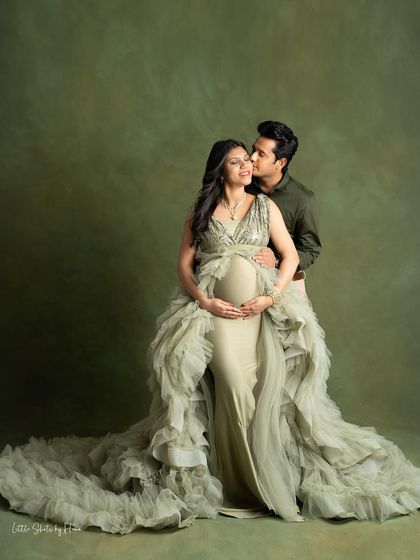 An elegant and romantic portrait of a couple against a textured olive green backdrop. The partner gently kisses the mother-to-be, who is wearing a stunning ruffled gown, showcasing a moment of pure affection.