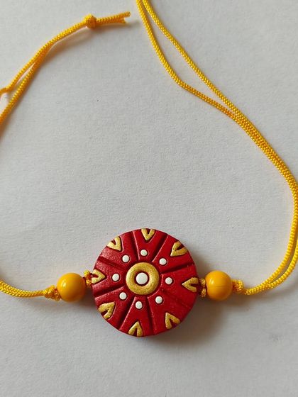A beautiful terracotta rakhi with a traditional red and gold design on a yellow thread. The intricate dots and patterns give it a festive and auspicious look.