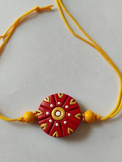 A beautiful terracotta rakhi with a traditional red and gold design on a yellow thread. The intricate dots and patterns give it a festive and auspicious look.