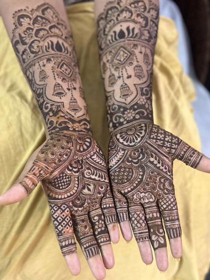 This traditional design features hanging jhumkas (bells) on the forearms and the bride's name 'Shina' hidden in the palm.