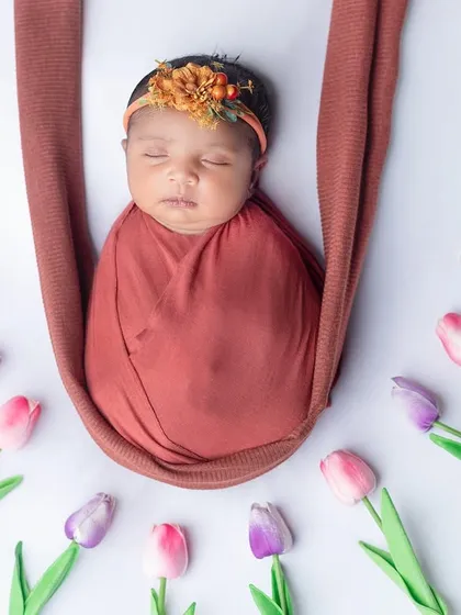 A newborn girl in a rust-colored wrap, hanging in a sling surrounded by tulips.