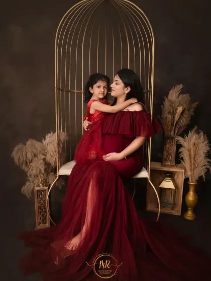 A loving mother and daughter portrait. The mother-to-be, in a deep red gown, sits in a golden birdcage chair while her daughter gives her a warm hug.