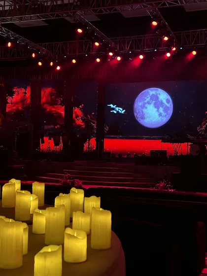 The stage is set for a performance, with a giant moon on the LED screen and a foreground of pillar candles, creating a moody and atmospheric setting.