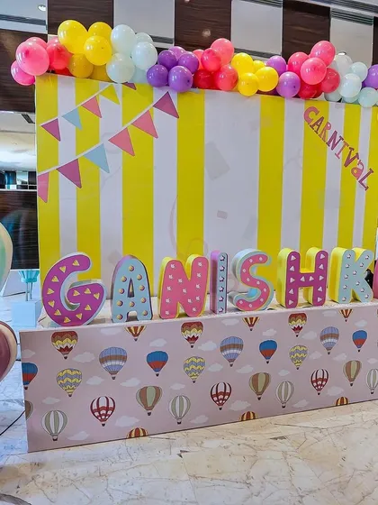 A personalized carnival name display for "Ganishka." The setup features large, colorfully decorated letters, a hot-air-balloon-themed table skirt, and a vibrant yellow-and-white striped backdrop topped with a balloon garland.
