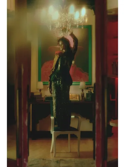 A dramatic wide shot where the model stands on a chair, an act of defiance and self-assurance. The styling is glamorous and unapologetic, marking the climax of her journey.