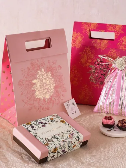 A festive gift bag in vibrant pink, perfect for Diwali. It includes a box of fudge brownies, a potli of honey choco almonds, and a box of sugar-free mithai.