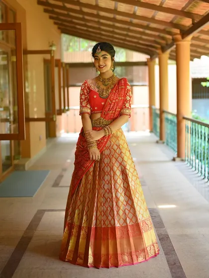 A celebration of classic bridal colors, this red and golden designer half saree is a masterpiece of bespoke beauty. The pure Kanchipuram silk lehenga is paired with a heavily embroidered red blouse, where every stitch tells a story of elegance and tradition, perfect for a South Indian bride.