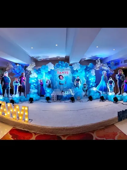 A panoramic view of a grand Frozen themed birthday stage. The entire setup is bathed in blue light, with character cutouts and balloon arrangements creating an icy, magical kingdom.
