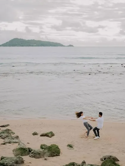A wide shot capturing the scale of the beach in Phuket, with Harman and Komal sharing a playful moment. The distant island adds to the beautiful scenery.