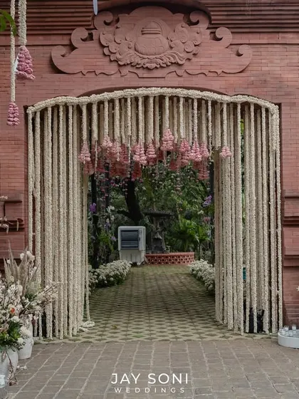 A beautiful fusion of traditional and rustic elements. This wedding entrance features a gate draped in tuberose garlands against a terracotta brick wall, flanked by arrangements of pampas grass and white florals in large urns.