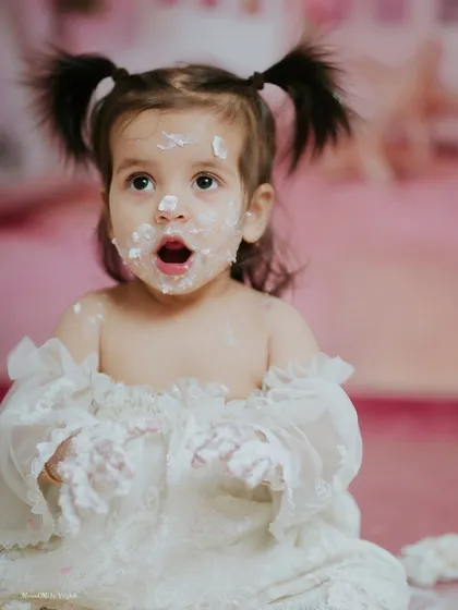 A look of pure wonder. The soft pink background and her delicate white dress create a dreamy atmosphere for her first birthday cake smash.