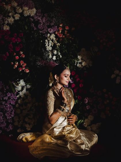 A dramatic portrait of a bride against a dark floral background. The lighting highlights the rich texture of her golden saree and the intricate details of her embroidered blouse.