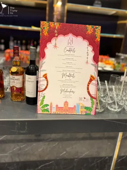A close-up of a custom wedding bar menu, designed with traditional Indian motifs to match the event's theme. Premium spirits like Glenlivet 15 are placed alongside, indicating the quality of our offerings.