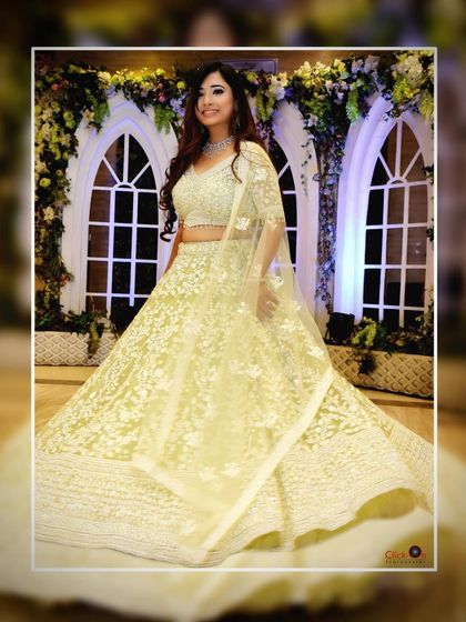 A full-length shot of the bride-to-be at her engagement, showcasing her beautiful white and gold lehenga against a floral backdrop.