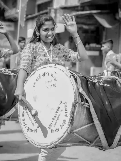 A black and white shot of a smiling female dhol player waving.