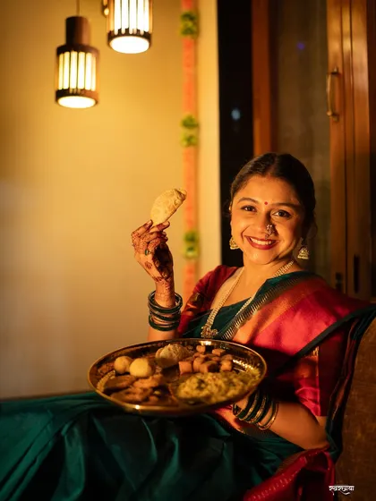 A joyful Diwali portrait showing the model holding a plate of traditional sweets ('faral'). Her bright smile and the warm indoor lighting create a feeling of home and celebration.