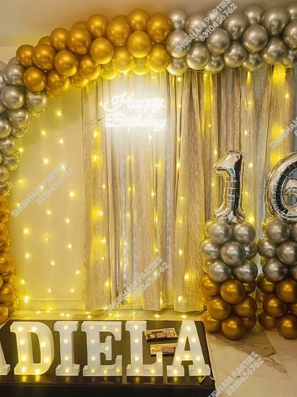 A chic 16th birthday party decoration with a gold and silver balloon arch, fairy light curtain, and marquee letters.