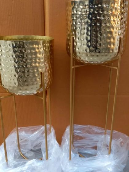 A pair of large, gold-finished metal planters with stands. The hammered texture on the pots adds a touch of handcrafted elegance, making them a statement piece for any living room or entryway.