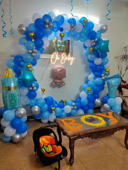 A "Welcome Baby Boy" setup in a living room, with a blue and white balloon ring, "Oh Baby" neon sign, and flower petal art on the table.