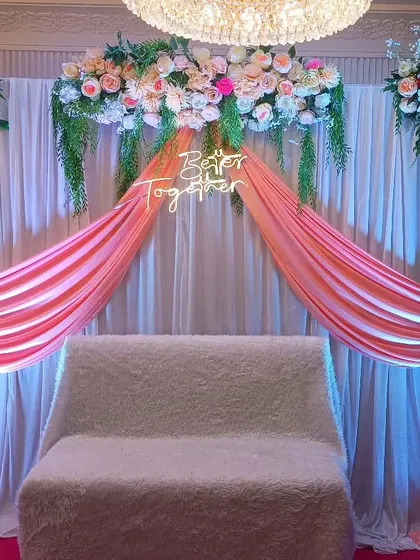 For this engagement, we used soft peach drapes and lush floral arrangements on the top and sides. The setup is illuminated with spotlights to highlight the stage area in a banquet hall.