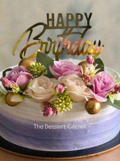 An elegant birthday cake with a soft purple ombre effect. The design is topped with a beautiful arrangement of fresh flowers and a gold "Happy Birthday" sign.
