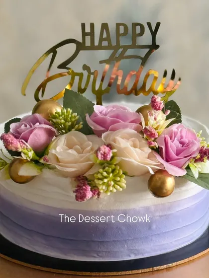 An elegant birthday cake with a soft purple ombre effect. The design is topped with a beautiful arrangement of fresh flowers and a gold "Happy Birthday" sign.