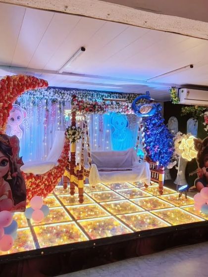 A beautiful stage setup for a naming ceremony, featuring a traditional jhula, a modern lighted moon prop, and cute character cutouts. This blend of elements caters to various tastes.