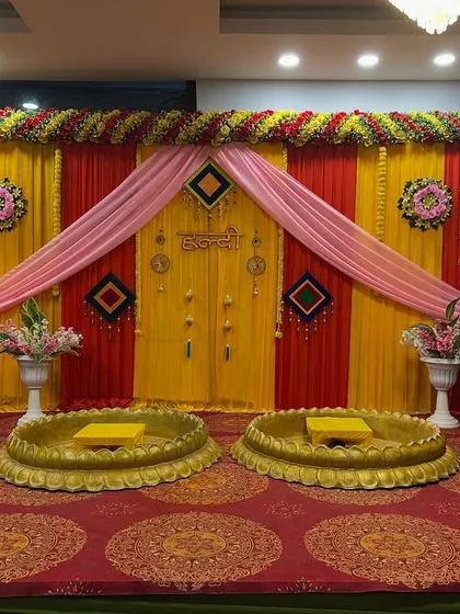 A colorful Haldi stage with red and yellow drapes, pink accents, and two lotus bowls for the bride and groom.