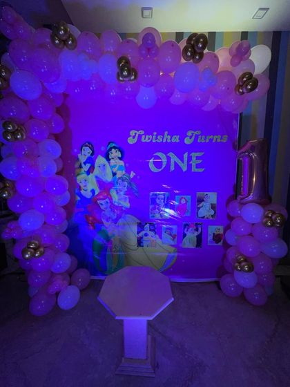 A Disney Princess theme for a first birthday. The pink backdrop features multiple princesses and is framed by a pink and gold balloon arch.