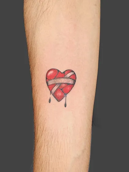 A small, broken heart tattoo held together by a bandage. This minimalist design uses a pop of red to convey deep emotion in a simple, artistic way.