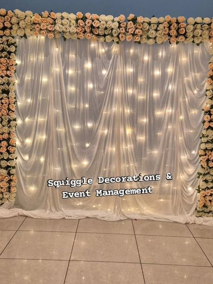 A simple yet elegant photo backdrop for any occasion. This setup features a curtain of fairy lights behind sheer white drapes, framed by a border of delicate artificial roses.