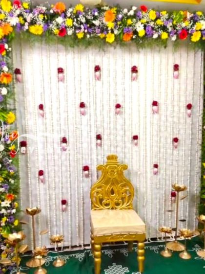 A beautiful floral frame backdrop made with a variety of colorful flowers. The center features a white curtain with hanging rose buds, and the setup is completed with a golden chair and brass lamps.