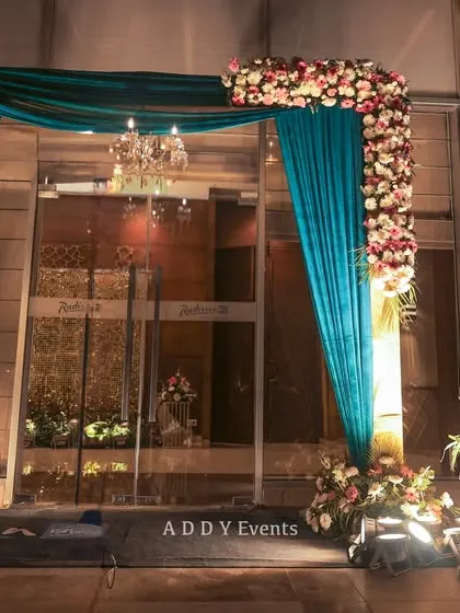 A beautiful floral and draped entrance gate for a Sangeet night, combining rich teal fabric with fresh flowers and a delicate chandelier.