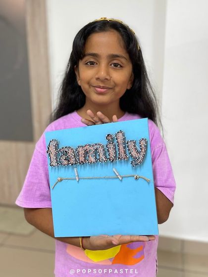 A young girl proudly shows off the "Family" string art she made, a heartfelt piece to display her favorite photos.