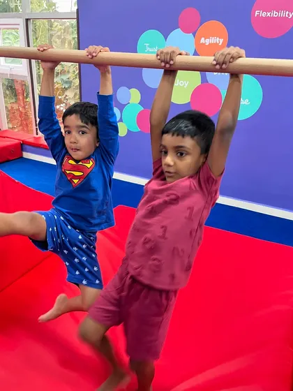Two boys, one in a Superman shirt, hang from the bar together. This activity builds strength and endurance, and doing it alongside a friend adds an element of fun and friendly competition.