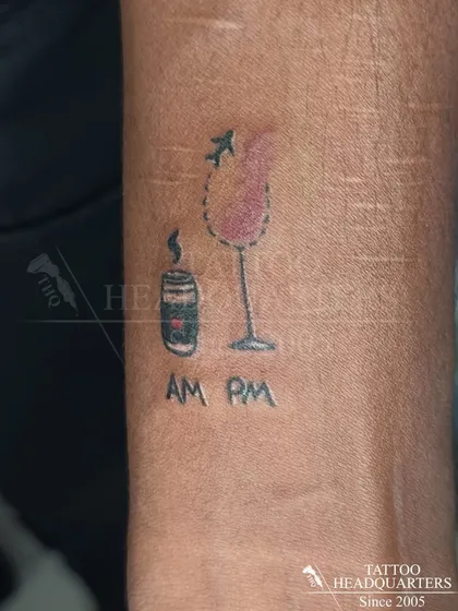 A quirky minimalist tattoo depicting a coffee cup for the "AM" and a wine glass for the "PM," symbolizing the daily routine.
