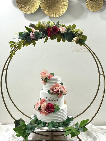 This three-tiered rustic wedding cake is displayed on a beautiful floral hoop stand, creating a stunning focal point. The cake features a textured whipped cream finish and is decorated with fresh, beautiful flowers.