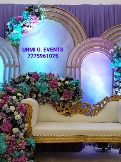I love creating these lush, floral-heavy designs for a truly romantic anniversary celebration. This stage features a cascade of purple, blue, and pink flowers flowing over an elegant white and gold sofa, set against backlit arched panels.
