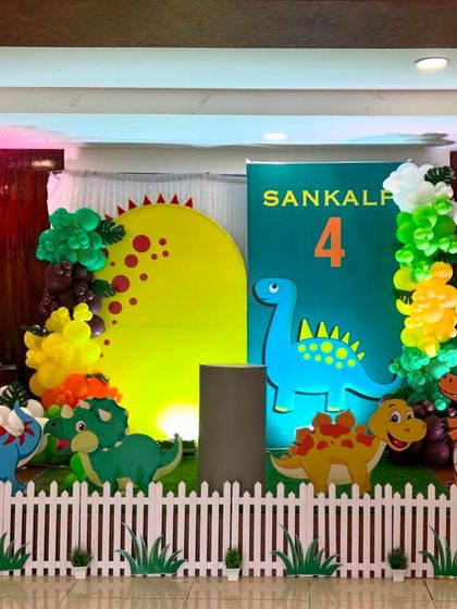 A friendly dinosaur theme for a 4th birthday party. The setup includes a white picket fence, cute cartoon dinosaur cutouts, and balloon arrangements in green and yellow to create a prehistoric jungle feel.