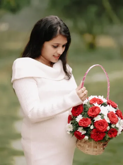 A serene portrait of an expectant mother in a classic white dress, holding a basket of vibrant red and white flowers. This image highlights a simple yet elegant prop that can add a touch of color and beauty to a maternity shoot.