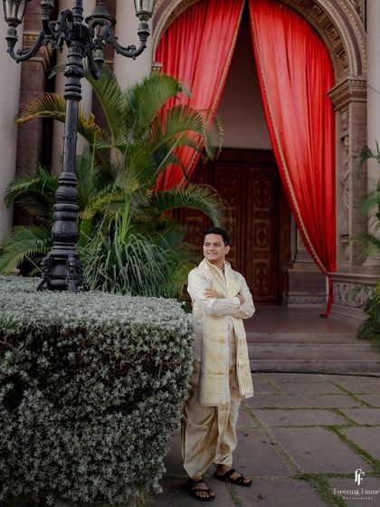 A portrait of the groom in his traditional dhoti, standing outside a grand venue.