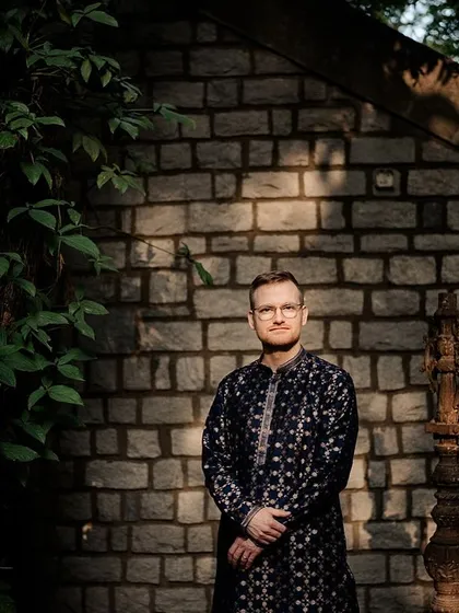 A groom in a navy blue kurta stands against a stone wall, the dappled sunlight creating a dramatic and striking portrait.