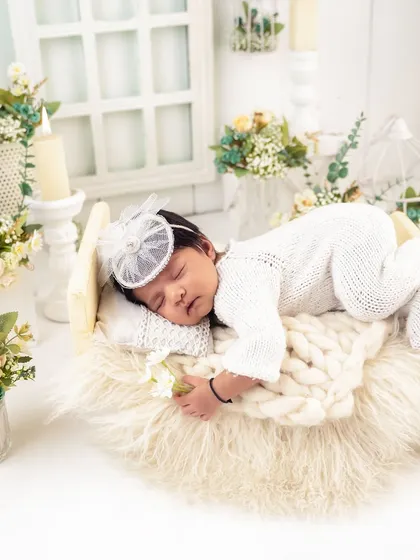 This peaceful portrait captures a baby sleeping on a miniature bed, holding a single white flower. The scene is filled with soft light and gentle floral details.