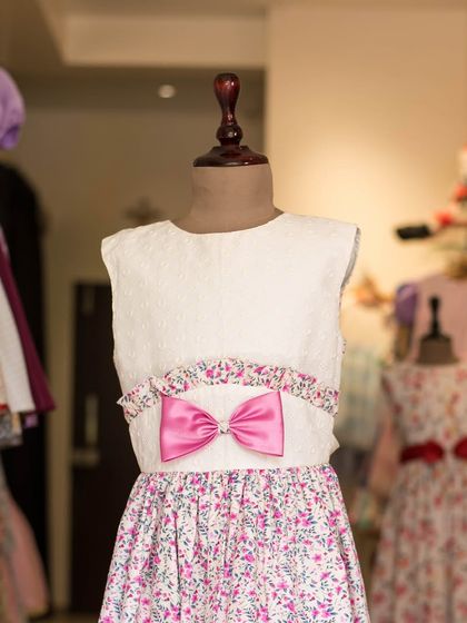 This dress combines a simple white bodice with a vibrant floral skirt. A bright pink bow at the waist adds a fun pop of color, tying the two parts of the dress together.