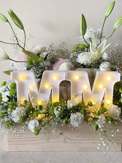 The beautiful white floral hamper itself, featuring the initials 'MM' in light-up letters surrounded by fresh lilies and chrysanthemums.