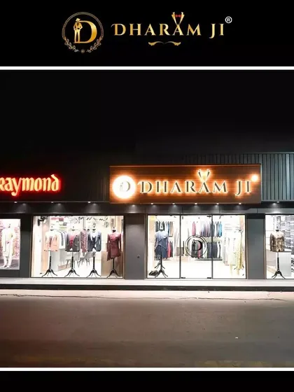 Our storefront at night, with the "Dharam Ji" and "Raymond" signs brightly lit. We are an authorized Raymond dealer, offering their quality fabrics alongside our own bespoke designs.