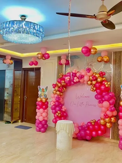 A grand and stylish "Welcome Our Little Princess" setup in a spacious living room. The centerpiece is a large pink backdrop surrounded by a ring of pink and gold balloons, flanked by fairy-themed balloon pillars.