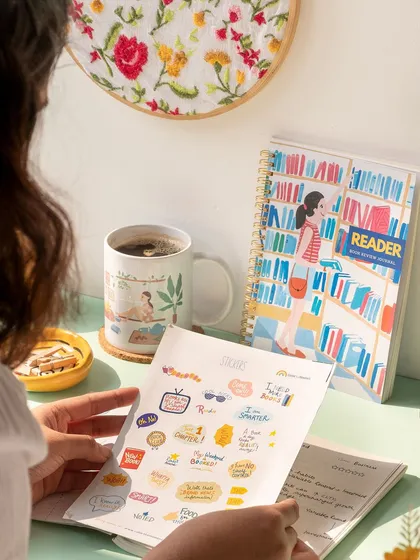 A cozy, reader-themed scene for a stationery brand. The over-the-shoulder perspective invites the viewer into the moment of journaling with a cup of coffee.