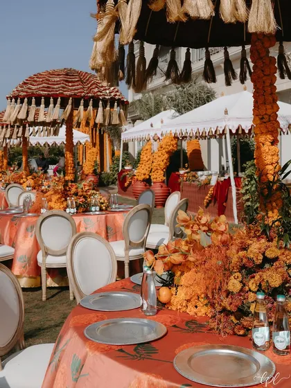 A vibrant Mehendi lunch setup with orange-themed tablescapes and large tasseled umbrellas. The use of bold, traditional colors and textures creates a festive and celebratory mood.